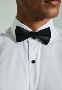 Papion texturat negru cu un nod central, purtat peste o cămașă albă cu guler, având un mic buton negru la gât.