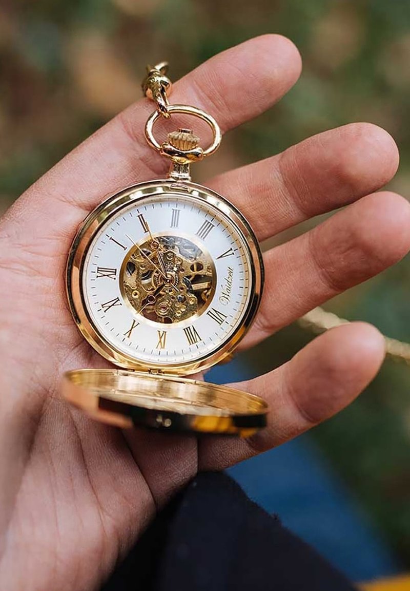 Goldene Taschenuhr mit einem weißen Zifferblatt, römischen Ziffern und einem Skelettdesign, das die inneren Abläufe zeigt. In einer Hand gehalten.