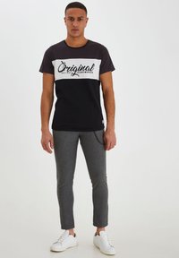 Schwarz-weißes Crew-Neck-T-Shirt mit „Original“-Druck, kombiniert mit grauen Nadelstreifen-Hosen und weißen Sneakern, stehend vor einem einfarbigen Hintergrund.