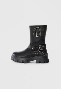 Bota de goma negra con hebillas y tachuelas plateadas, acabado en cuero texturizado, suela gruesa y detalle de cadena en el lateral.