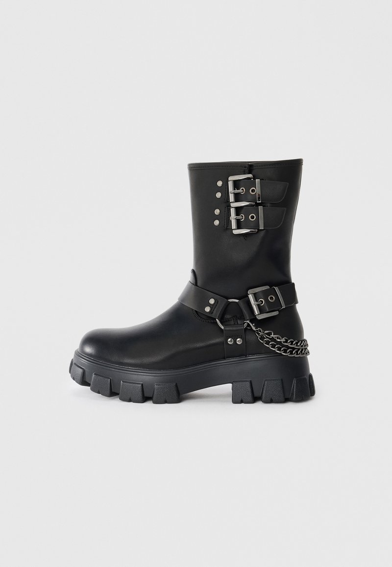 Bota de goma negra con hebillas y tachuelas plateadas, acabado en cuero texturizado, suela gruesa y detalle de cadena en el lateral.