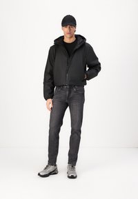 Črna zip-up jopica s kapuco, nosena preko črne majice; v kombinaciji s temno sivimi kavbojkami in sivimi čevlji. Model nosi ujemajočo se kapo.