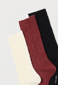 Τρία ζευγάρια ριγέ πλεκτών κάλτσων σε κρέμα, μπορντώ και μαύρο, τοποθετημένα το ένα πάνω στο άλλο σε ανοιχτό φόντο.