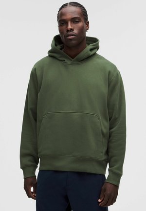 Hombre con el pelo trenzado, vestido con sudadera con capucha verde oliva y pantalones oscuros, de pie frente a un fondo claro y liso.