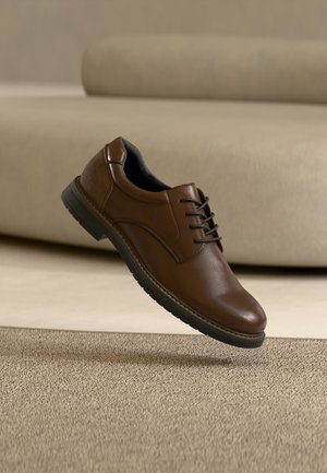 Brun læderdressko med sort sål og snørebånd svæver tilsyneladende over et beige tæppe i et blødt oplyst, moderne indendørs miljø.