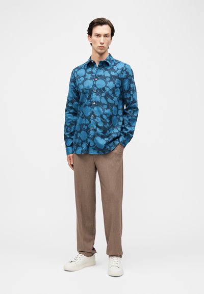 Άνδρας στέκεται φορώντας μπλε πουκάμισο με λουλουδάτο μοτίβο και κουμπιά, μπεζ παντελόνι και λευκά αθλητικά παπούτσια, μπροστά σε απλό φωτεινό φόντο.