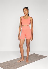 Korallfarget treningsantrekk som inkluderer en tettsittende crop-top og høyt liv shorts, laget av glatt stoff. Stående på en teksturert yogamatte.