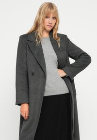 Abrigo gris de mezcla de lana que presenta un diseño de doble botonadura, grandes solapas con muescas y una silueta relajada. Se combina con un suéter gris claro.