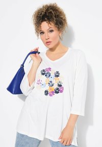 Weißes langärmliges T-Shirt mit V-Ausschnitt und mehrfarbigem Blumenmuster sowie dem Text "CASUAL MODE", kombiniert mit einer blauen Handtasche. Sichtbare Jeans aus Denim.