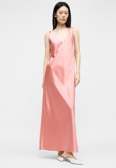 Γυναίκα με αμάνικο, μεταξωτό, ανοιχτό ροζ φόρεμα έως το πάτωμα με βαθύ V ντεκολτέ, συνδυασμένο με λευκά μυτερά τακούνια, στέκεται σε λευκό φόντο.