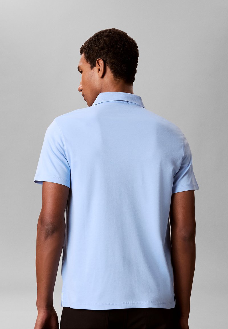 Man met zijn rug naar de camera, draagt een lichtblauw poloshirt met korte mouwen tegen een effen grijze achtergrond.