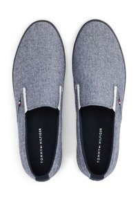 Ett par blå slip-on sneakers med vita sulor och Tommy Hilfiger-logotyp inuti, visade ovanifrån på en vit bakgrund.
