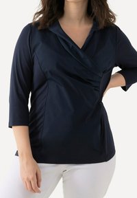 Top blu navy con colletto scialle, maniche a tre quarti e arricciature laterali. Texture liscia, design aderente e orlo curvo.