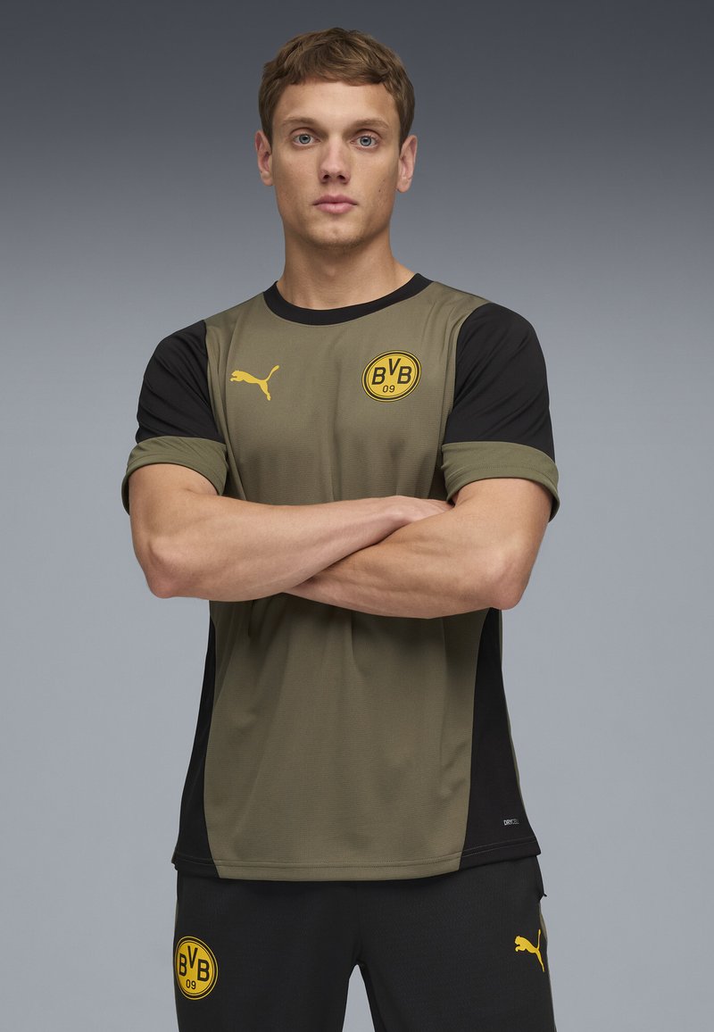 Puma sportsjersey i oliven grøn og sort, med korte ærmer, der har et cirkulært gult logo og en sort detalje på ærmerne. Glat tekstur.