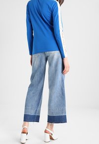 Blå långärmad skjorta med vita sidostreck, kombinerad med ljusblå vida jeans med fransad nederkant och kontrasterande denimpanel. Vita klacksandaler.