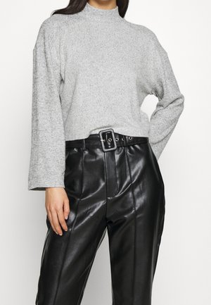 Vrouw draagt een lichtgrijze gebreide trui met lange mouwen, ingestopt in hooggetailleerde zwarte leren broek met een brede riem en een vierkante gesp.