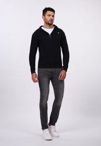 Schwarze Zip-Hoodie mit Kängurutasche, kombiniert mit grauen Slim-Fit-Jeans und weißen Sneakers. Mit Kordelzügen und kleinem Logo.