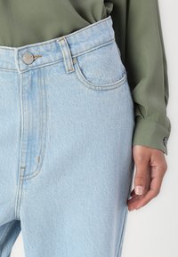 Lyseblå denimjeans har høy midje, synlig søm, frontlommer og en enkel metallknapp. En grønn bluse vises delvis.