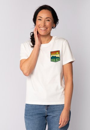 T-shirt en coton blanc avec un patch à l'avant représentant une scène de montagne colorée, un soleil et le texte "GOOD VIBE". Coupe décontractée, manches courtes.