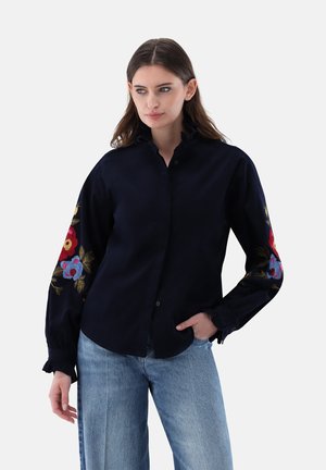 Frau trägt ein schwarzes Hemd mit bunter Blumenstickerei an den Ärmeln und hellblaue Jeans, steht mit einer Hand in der Tasche.