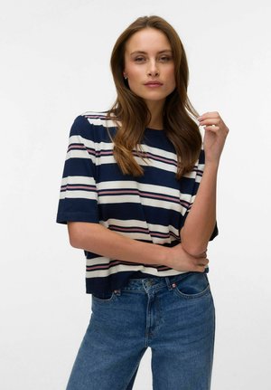 Mlada žena s dugom smeđom kosom nosi majicu na pruge u mornarsko plavoj, bijeloj i ružičastoj boji s kratkim rukavima te plave traperice, stojeći ispred jednobojne pozadine.