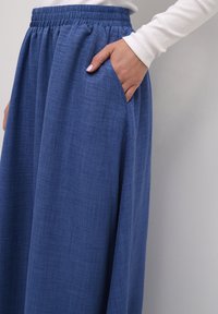 Blå stofnederdel med en struktureret overflade, der har en elastisk talje og sidelommer. Stofmaterialet hænger løst omkring benene.