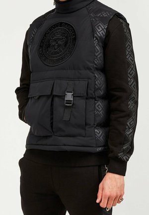 Uomo che indossa un gilet imbottito nero con grande emblema ricamato di ape e tasca frontale con fibbia, sopra una camicia nera a maniche lunghe con motivo a fantasia.