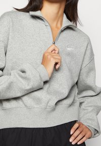 Grå, zip hoodie gjord av mjuk bomullsblandning. Har en hög krage, ribbade manschetter och nederkant, med en liten vit Nike-logga på bröstet.