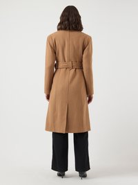 Long brown coat made of soft wool, featuring a belted waist, straight cut, and seam detailing. Worn over black pants and heeled shoes.