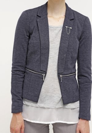 Personne portant un blazer gris foncé avec des poches zippées et une broche en argent sur un haut gris clair en couches et un pantalon clair.