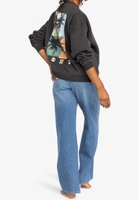 Persona che indossa jeans larghi blu e una felpa scura con grafiche di palme sulla schiena, in piedi a piedi nudi su uno sfondo bianco.