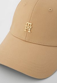 Beige baseballpet met een gestructureerd ontwerp en een glanzend gouden logo aan de voorkant. De stof heeft een ribbeltextuur en zichtbare stiksels.