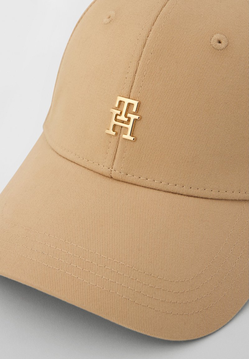 Beige baseballpet met een gestructureerd ontwerp en een glanzend gouden logo aan de voorkant. De stof heeft een ribbeltextuur en zichtbare stiksels.