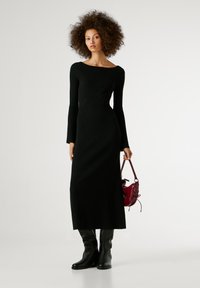 Juoda raštuota megztinė su ilgomis plačiomis rankovėmis, valtimi pjaustyta iškirpte ir aptempta siluetu; dėvima su juodais aukštakulniais batais iki kelių ir burgundiška rankine.