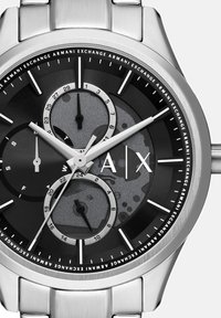 Hõbedane roostevabast terasest kell, millel on must ja hall dial, kolm alade pakkumist, luminofoorsed tunni markerid ja logo kell 3.