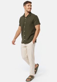Camicia verde scuro a maniche corte con un design testurizzato, bottoni bianchi, pantaloni beige e sandali scuri dallo stile minimalista.