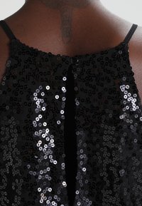 Vêtement noir orné de sequins, avec un décolleté rond, des bretelles fines et une fermeture à boutons au dos avec une découpe en forme de goutte d'eau.