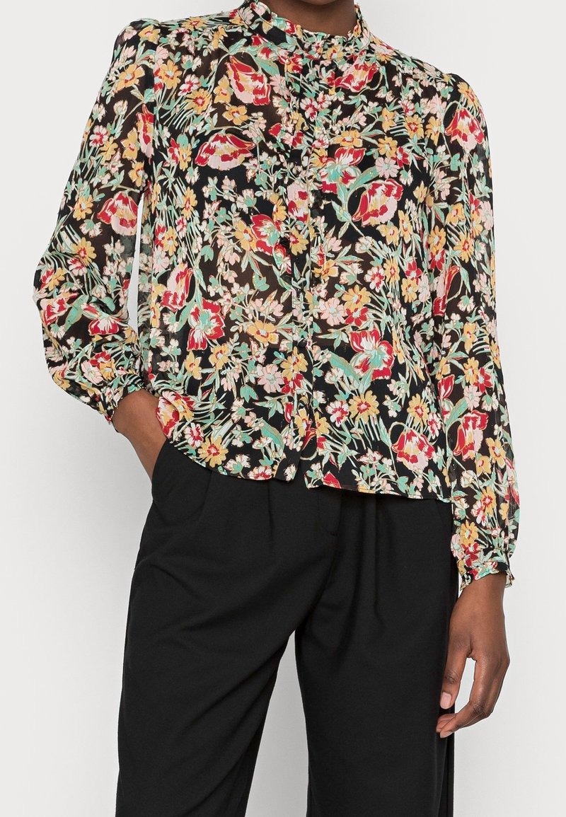 Blouse transparente à imprimé floral avec manches longues bouffantes, associée à un pantalon noir ample, main posée négligemment dans la poche.