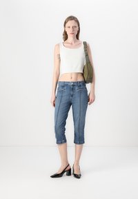 Débardeur blanc court, capris en denim bleu avec deux poches avant et des surpiqûres contrastées, et des chaussures à talons noirs. Sac à bandoulière vert olive.
