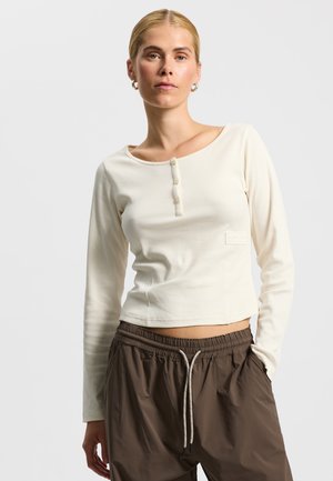 Langærmet ribbet top i off-white med rund halsudskæring, tre-knaps lukning og et diskret logopatch på siden.