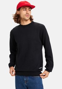 Black sweater with long sleeves, round neckline, and ribbed cuffs, paired with blue jeans. Features a white logo detail on the hem.