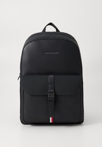 Sac à dos noir en matériau synthétique lisse. Il possède un rabat à l'avant avec une boucle, un logo ton sur ton et un accent rayé en bas.