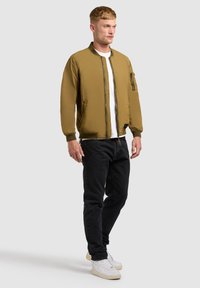 Jeune homme debout, portant une veste aviateur beige, un t-shirt blanc, un jean noir et des baskets blanches, sur un fond gris uni.