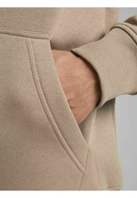 Sudadera beige con bolsillo tipo canguro, puños acanalados y textura suave. Presenta costuras contrastantes a lo largo del borde del bolsillo.