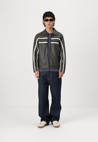 Black leather jacket with white stripes, purple shirt underneath, loose dark denim jeans, and white shoes; model wearing dark sunglasses.