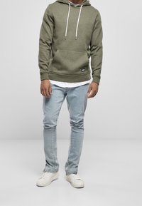 Olivgrüner Hoodie mit einer Fronttasche, weißen Kordeln und gerippten Bündchen, getragen über einem weißen Hemd; kombiniert mit hellblauen Jeans und weißen Schuhen.