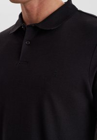 Svart pikétröja med en texturerad tyg, två knappar vid kragen och en subtil logotyp på vänster bröstområde. Korta ärmar.