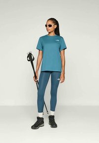 Türkiz sportpóló és leggings, fekete túracipő, trekkingbotot tart. Napbarnító és logó részlet a ruházaton. Simább textúra.