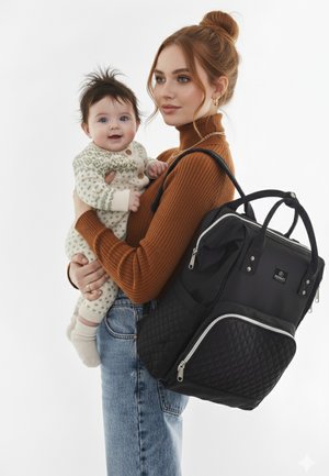 Sort quilted rygsæk med sølv lynlåse, båret mens en baby i en mønstret cremefarvet natdragt holdes. Baggrunden er ren hvid.