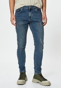 Hombre con jeans ajustados azul desgastados y zapatillas verde de caña alta con suelas gruesas beige, de pie contra un fondo blanco.